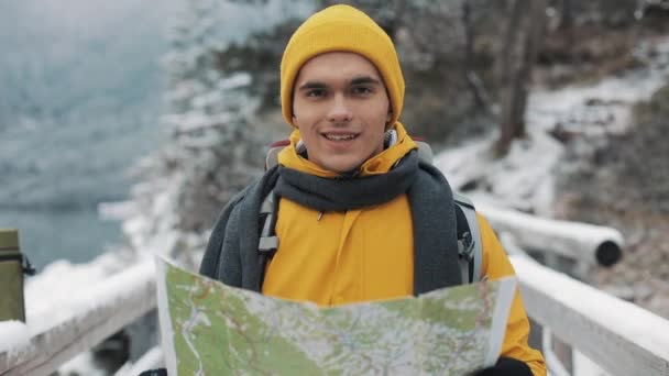 Ung attraktiv mann med gule vinterklær som holder kart og ser inn i kameraet. Portrett av en turist som står på broen. Vakre fjell om vinteren i bakgrunnen – stockvideo