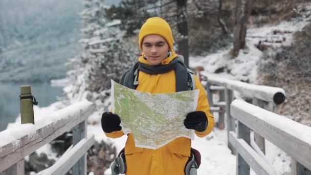 Ung attraktiv mann med gule vinterklær som holder kart og ser inn i kameraet. Portrett av en turist som står på broen. Vakre fjell om vinteren i bakgrunnen – stockvideo