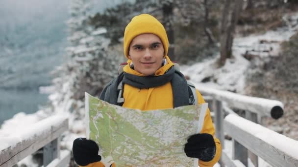 Молодой привлекательный мужчина в желтой зимней одежде держит карту и смотрит в камеру. Портрет туриста, стоящего на мосту. Красивые горы в зимнее время на заднем плане — стоковое видео