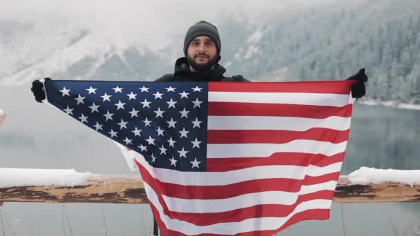 Мандрівник чоловік тримає прапор Америці стоїть у горах Сніг накривав акуратні beautuful озера. Він дивиться в камеру і посміхається. Мандрівного мандрівник — стокове відео