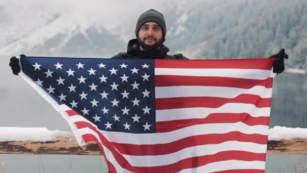 Gezgin adam Amerika ayakta bayrağı tutarak karla kaplı dağlara düzgün beautuful Gölü. Kameraya bakıyor ve gülümsüyor. Uzun yürüyüşe çıkan kimse gezgin — Stok video