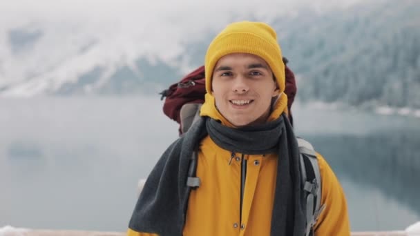 冬服の魅力的な若者の肖像画 ハイキング男黄色の冬の服を着てはカメラと笑顔に見えます 美しい山々 と雪に覆われた湖の背景 — ストック動画