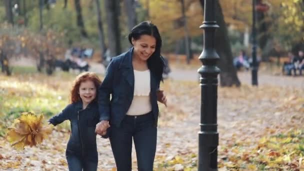 Feliz joven madre y su pequeña hija pelirroja corriendo juntas en un parque de otoño. Se ríen y se divierten tomados de la mano. Movimiento lento — Vídeo de stock
