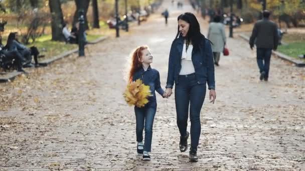 A beautiful mother and her cute little daughter walking in the autumn park. They walk around in the middle of people. Little girl holding yellow leaf bunch in her hand — Stock Video