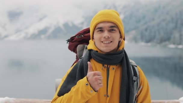 Porträtt av en ung vandring man med ryggsäck stående stående på naturen på vintern, ger tummen upp. Vackra berg och snötäckta sjön bakgrund — Stockvideo