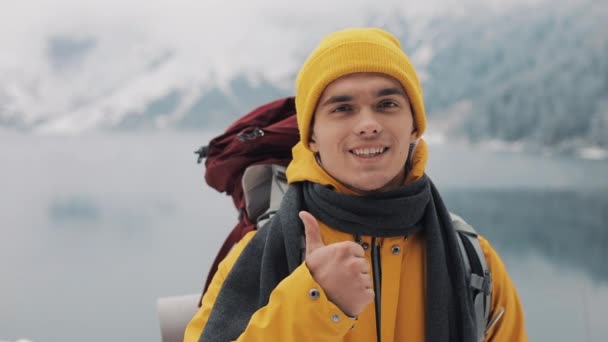 Portréja egy gyalogos fiatalember hátizsák állandó állandó jellegű télen, hüvelykujj feladom. A gyönyörű hegyekkel és a hóval borított tó háttér — Stock videók