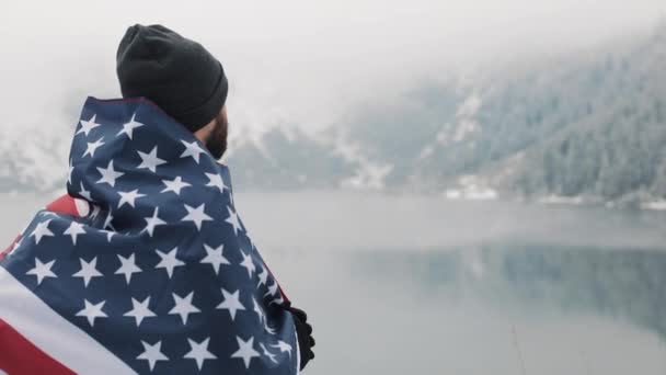 美しい湖の近く雪に覆われた山の中のアメリカの地位のフラグと旅行者の男。美しい自然を見てハイカー — ストック動画