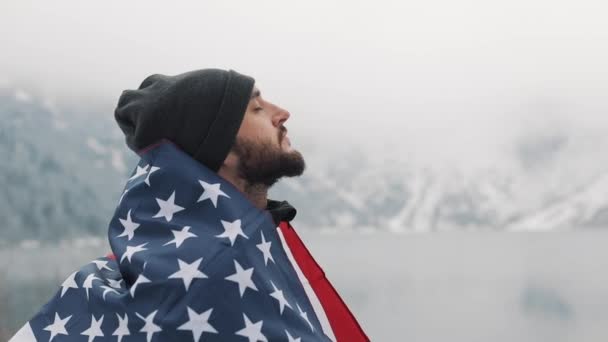 Homem viajante com bandeira da América em pé nas montanhas cobertas de neve perto de belo lago. Caminhante olhando para a bela natureza — Vídeo de Stock