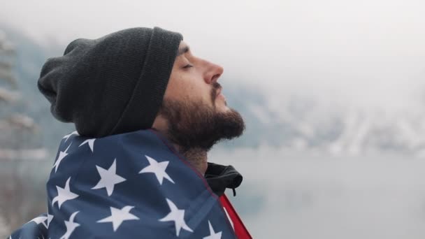 아름 다운 호수 근처의 눈 덮인 산에 서 있는 미국 국기와 함께 여행자 남자. 아름 다운 자연에서 찾는 등산객 — 비디오