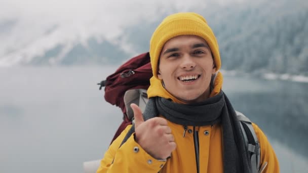親指をあきらめるとバックパック、冬の自然に立っている立っている若いハイキングの男の肖像画。美しい山々 と雪に覆われた湖の背景 — ストック動画