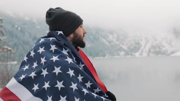 Hombre viajero con bandera de América de pie en las montañas cubiertas de nieve cerca de un hermoso lago. Caminante mirando la hermosa naturaleza — Vídeos de Stock