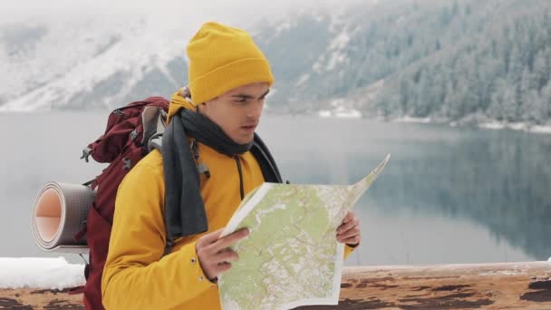 Viajar nas montanhas. O jovem caminhante usa um mapa em papel da área. Procura o caminho. Gosta de aventura e viagens. Em um fundo de montanhas de inverno e um belo lago — Vídeo de Stock