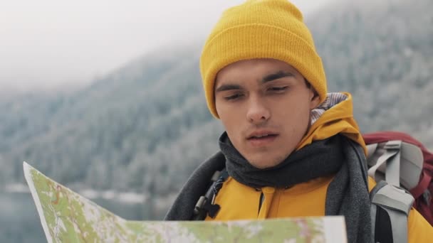 Путешествие по горам. Молодой турист использует бумажную карту местности. Ищет путь. Любит приключения и путешествия. На фоне зимних гор и красивого озера — стоковое видео