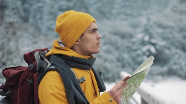 Joven hombre atractivo vistiendo ropa de invierno amarilla sosteniendo el mapa de pie en el puente y revisando la ruta. Hermosas montañas en invierno en el fondo — Vídeo de stock