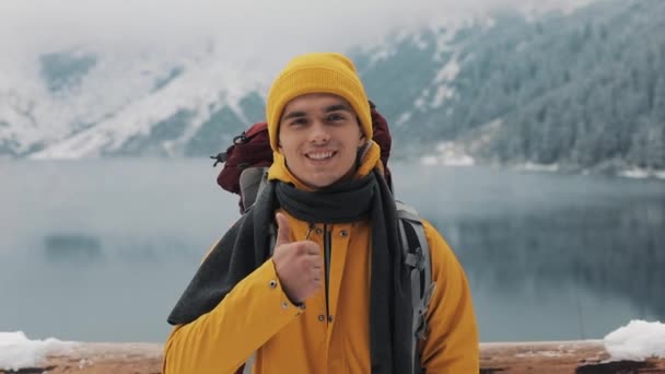 초상화는 하이킹 젊은이 배낭 서 서 자연에는 겨울에 엄지손가락을 포기. 아름 다운 산과 눈 덮인 호수 배경 — 비디오