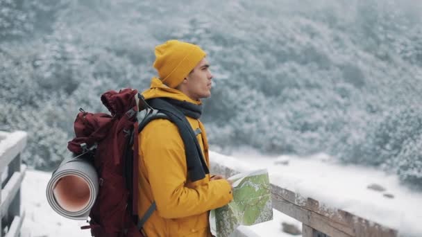 Joven hombre atractivo vistiendo ropa de invierno amarilla sosteniendo el mapa de pie en el puente y revisando la ruta. Hermosas montañas en invierno en el fondo — Vídeos de Stock