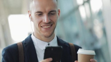 Smartphone kahve içme ve kulaklık ile kullanarak ofis binası yakınında duran genç iş adamı. İş adamı moda siyah takım elbise