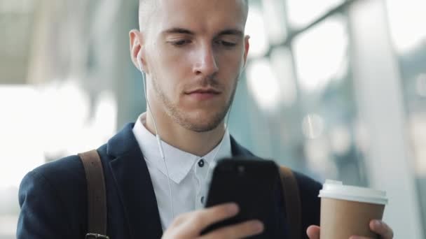 Giovane uomo d'affari in piedi vicino a palazzo degli uffici utilizzando smartphone con cuffie e bere caffè. Uomo d'affari in abito nero moda — Video Stock