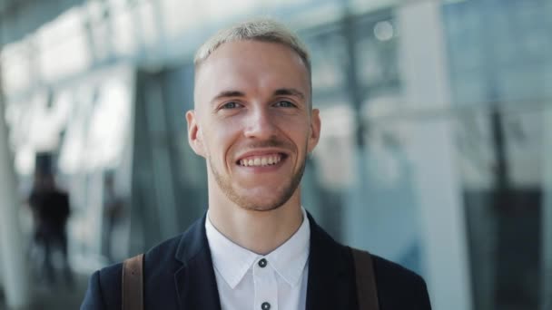 Портрет усміхненого молодого бізнесмена з рюкзаком стоячи поза аеропортом і дивлячись в камеру — стокове відео
