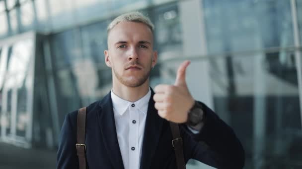 Homem de negócios atraente jovem que está perto do aeroporto e olhando para a câmera. Homem mostra polegar para cima e para baixo — Vídeo de Stock