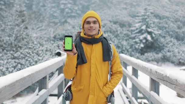 Retrato de un joven atractivo en ropa de invierno que muestra el teléfono inteligente con pantalla verde mirando a la cámara. Chroma Key gadget naturaleza invierno — Vídeos de Stock