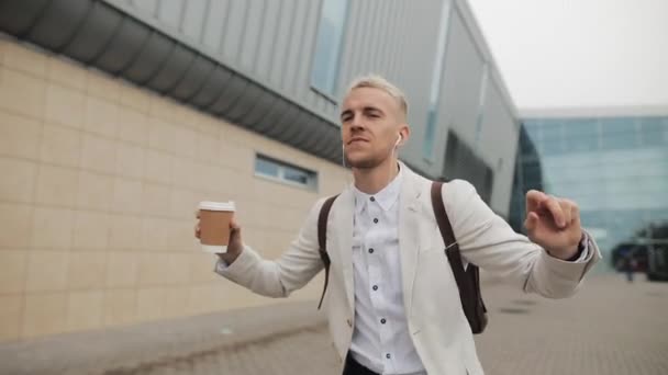 Šťastný mladý podnikatel s kávou poslechu hudby ve sluchátkách na smartphone a vtipný tanec, chůze nedaleko budovy úřadu. Zábavné taneční pohyby. Zpomalený pohyb — Stock video
