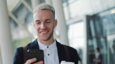 Sokakta yürürken ve smartphone kullanarak mutlu gülümseyen çekici bir adam. Git ve okuma iyi haber için kahve içme. Havalimanı arka plan üzerinde terminal