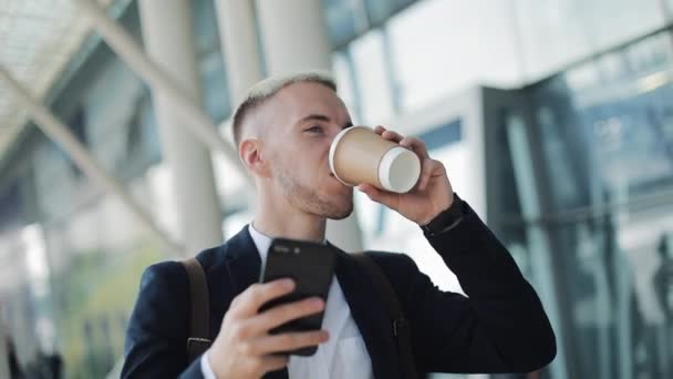 愉快的微笑有魅力的男人走在大街上 使用智能手机 他喝咖啡去看好消息 背景上的机场航站楼 — 图库视频影像
