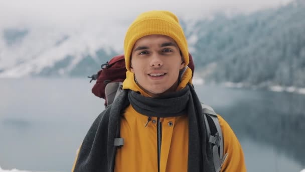 Porträtt av en ung attraktiv man i vinterkläder. Vandring man klädd i gul vinterkläder ser på kameran och leenden. Vackra berg och snötäckta sjön bakgrund — Stockvideo