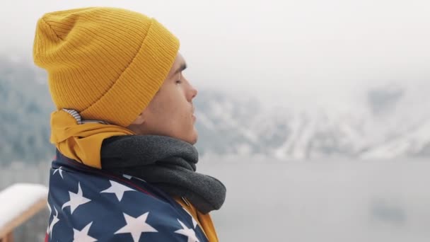 Hombre viajero con bandera de América de pie en las montañas cubiertas de nieve cerca de un hermoso lago. Caminante mirando la hermosa naturaleza — Vídeos de Stock