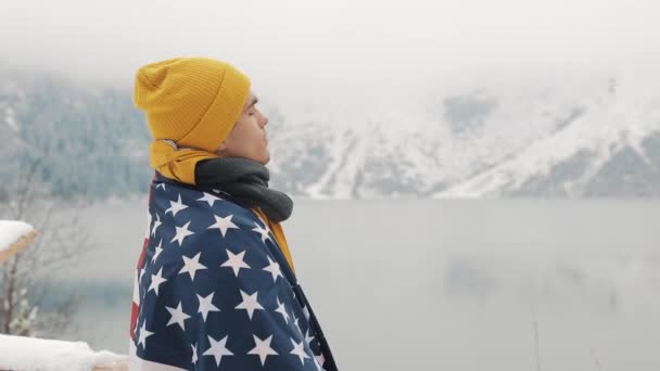 Homem viajante com bandeira da América em pé nas montanhas cobertas de neve perto de belo lago. Caminhante olhando para a bela natureza — Vídeo de Stock