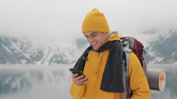 Giovane viaggiatore uomo utilizzando smartphone in escursioni tour invernale. Montagne innevate sfocate, promontorio e lago. Concetto di viaggio e comunicazione: scorrimento escursionistico e intercettazione on line tramite cellulare — Video Stock