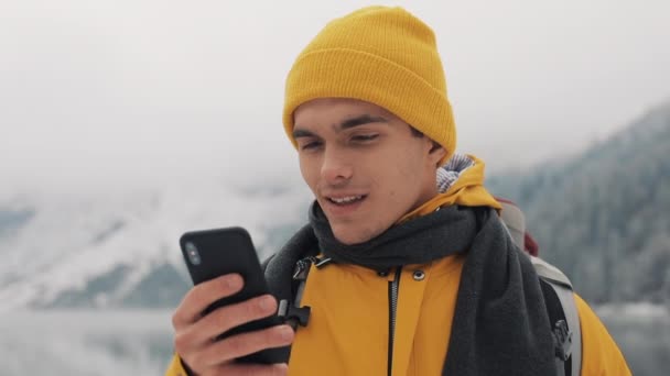 Jeune voyageur qui utilise son smartphone pour une randonnée hivernale. Montagnes enneigées floues cap et lac. Concept de voyage et de communication : défilement des randonneurs et écoute en ligne par téléphone portable — Video