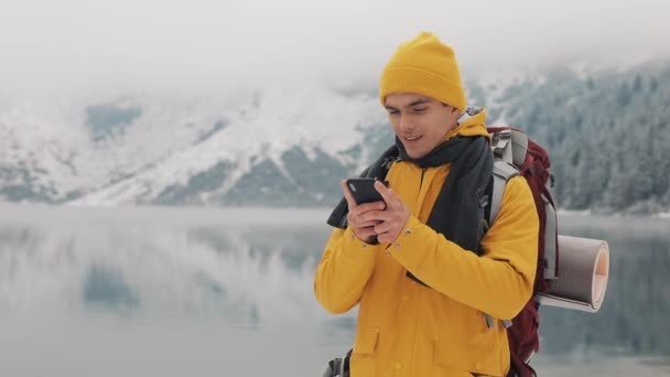 Człowiek młody podróżnik za pomocą smartfona w pieszej wędrówki zimowe. Przylądek Snowy góry niewyraźne i jezioro. Podróży i komunikacja koncepcja: turysta przewijanie i naciskając na linii przez telefon komórkowy — Wideo stockowe