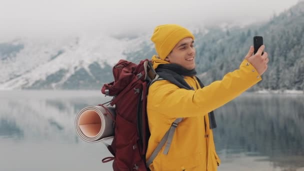 Красивые горы зимой. Человек с бородой, одетый в желтую зимнюю одежду, делает селфи на фоне красивых гор и горного озера — стоковое видео