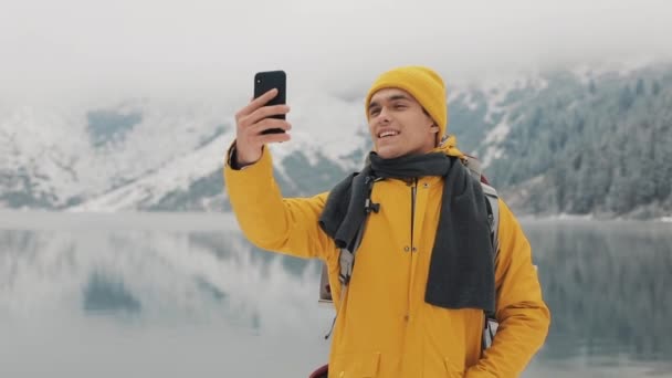 Красивий чоловік-поводир відео дзвінки на смартфон, махаючи на камеру, посміхаючись і розмовляючи з друзями. Прекрасні зимові гори і озеро на фоні, повільний рух — стокове відео