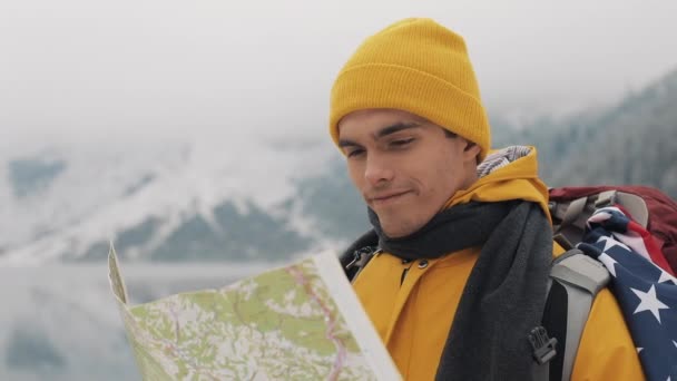 Viajando por las montañas. El joven excursionista americano utiliza un mapa en papel de la zona. Busca en el camino. Disfruta de la aventura y los viajes. Sobre un fondo de montañas de invierno y un hermoso lago — Vídeo de stock