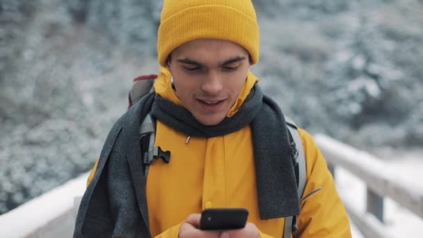 A young tourist standing on a bridge in the snowy mountains with a smartphone in his hands. Communication, social networks, travel concept — Stock Video