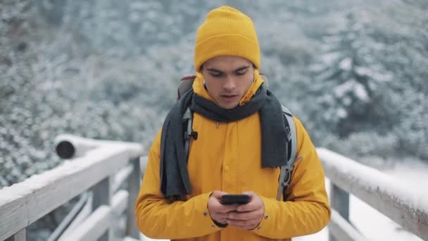 A young tourist walks on a bridge in the snowy mountains with a smartphone in his hands. Communication, social networks, travel concept — Stock Video