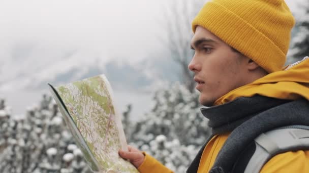 在山里旅行。身穿黄色冬衣的年轻人拿着地图站在被雪覆盖的树林里。旅行, 冒险, 攀岩 — 图库视频影像