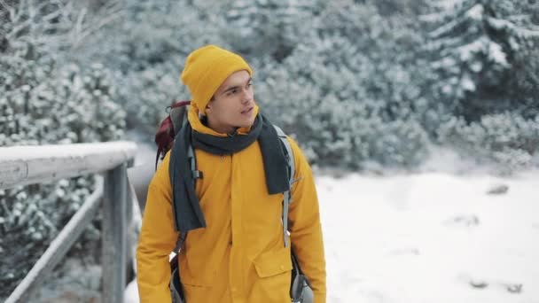 Voyager dans les montagnes. Jeune homme portant des vêtements d'hiver jaunes marchant sur la montagne recouverte de neige. Il va sur un pont à travers la rivière et regarde autour de lui. Voyage, aventure, escalade — Video