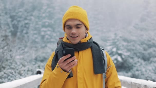 彼の手でスマート フォンでの雪の山の橋の上に立って 若い観光客 コミュニケーション 社会的ネットワーク 旅行のコンセプト — ストック動画