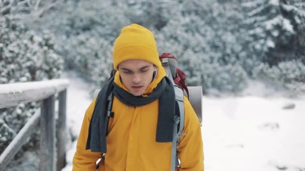 在山里旅行。身穿黄色冬衣的年轻人走在被雪覆盖的山上。他走在河对岸的一座桥上环顾四周。旅行, 冒险, 攀岩 — 图库视频影像