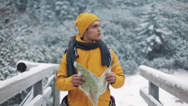 Подорожуючи в гори. Молодий чоловік у жовтому зимовому одязі тримає карту, що йде на гору, вкриту снігом. Поїздка, пригоди, скелелазіння — стокове відео