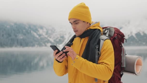 Hiker mannen med smartphone som tar ett foto av vinter natur. Turisten beundrar naturen och gör bilder. Ung man stående mot bakgrund av vackra berg — Stockvideo