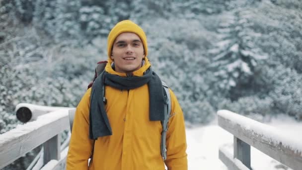 Viajar nas montanhas. Jovem vestindo roupas amarelas de inverno andando na montanha coberta de neve. Ele vai em uma ponte do outro lado do rio e olha para a câmera. Viagem, aventura, escalada — Vídeo de Stock