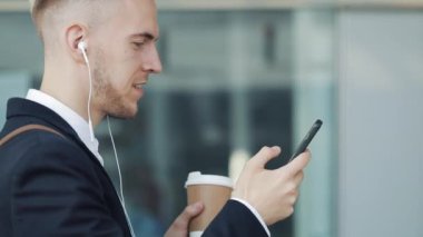 Genç çekici insan kahve tutarak ve telefon kullanarak sokakta yürüyor. İş Merkezi etrafında yürüyüş kulaklık müzik dinlediği