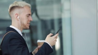 Genç çekici insan kahve tutarak ve telefon kullanarak sokakta yürüyor. İş Merkezi etrafında yürüyüş kulaklık müzik dinlediği