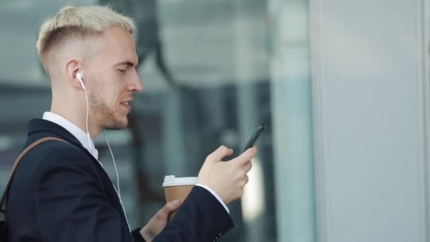 젊은 매력적인 남자는 커피를 들고와 휴대 전화를 사용 하 여 거리를 산책. 그는 비즈니스 센터의 주위에 산책 하는 헤드폰에서 음악을 듣으십시오 — 비디오