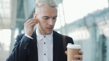 Genç çekici adam sokakta kahve tutarak yürür ve iş merkezi etrafında yürüyüş kulaklık müzik dinle